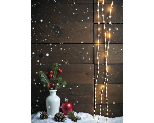 Weihnachtliche Dekoration mit LED-Lichterketten vor einer Holzwand, Vase mit Tannenzweigen, Beeren, Zapfen und einer Weihnachtskugel im Schnee.