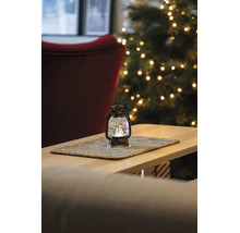 Weihnachtliche Dekolaterne mit Schneemann-Schneekugel auf einem Holztisch vor einem beleuchteten Weihnachtsbaum.