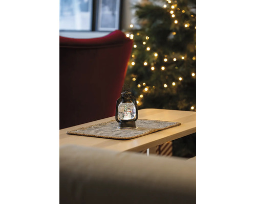 Weihnachtliche Dekolaterne mit Schneemann-Schneekugel auf einem Holztisch vor einem beleuchteten Weihnachtsbaum.