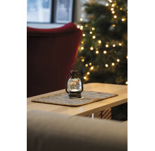 Beleuchtete LED-Wasserlaterne mit Rotkehlchen-Motiv auf einem Tischset in einem hellen Raum mit geschmücktem Weihnachtsbaum im Hintergrund.