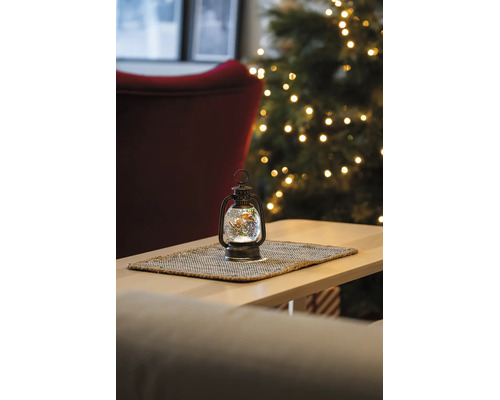 Beleuchtete LED-Wasserlaterne mit Rotkehlchen-Motiv auf einem Tischset in einem hellen Raum mit geschmücktem Weihnachtsbaum im Hintergrund.