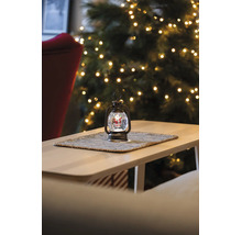 Weihnachts-Laterne mit Schneekugel und Weihnachtsmann-Figur auf einem Tisch vor einem beleuchteten Tannenbaum.