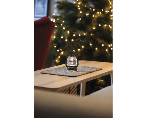 Weihnachts-Laterne mit Schneekugel und Weihnachtsmann-Figur auf einem Tisch vor einem beleuchteten Tannenbaum.