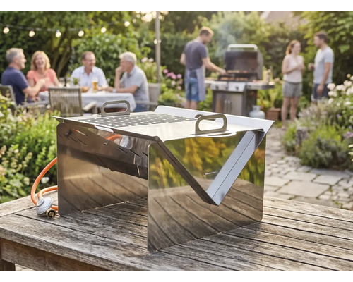 Tisch-Gasgrill aus Edelstahl auf einem Holztisch im Garten, im Hintergrund eine gesellige Runde von Personen beim Grillfest.