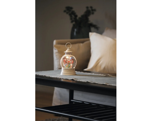 Lanterne boule à neige blanche lumineuse avec grande roue et sapin de Noël sur une table dans un salon clair et ordonné.