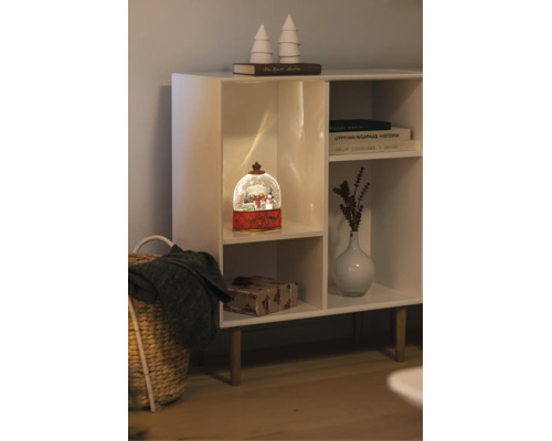 Beleuchtete Schneekugel mit Winterszene in einem weißen Regal neben einer Vase, Büchern und einem Geschenkpaket.