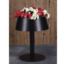 Schwarzer Pflanzkübel in Form einer Stehlampe mit roten und weißen Blumen auf grauem Boden vor einer Holzwand.
