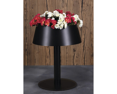 Schwarzer Pflanzkübel in Form einer Stehlampe mit roten und weißen Blumen auf grauem Boden vor einer Holzwand.