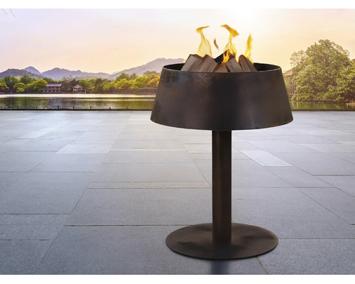 Moderne Feuerschale aus dunklem Metall auf einem Standfuß mit brennendem Holz auf einer Terrasse an einem See.