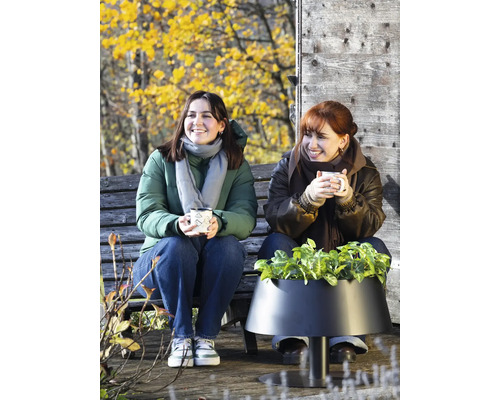 Zwei Frauen sitzen auf einer Holzbank neben einem modernen schwarzen Pflanzgefäß mit Grünpflanzen in einer herbstlichen Außenanlage.