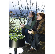 Zwei Frauen sitzen im Freien auf einer Holzterrasse neben einem modernen schwarzen Pflanzkübel mit grünen Pflanzen.
