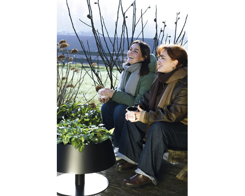 Zwei Frauen sitzen im Freien auf einer Holzterrasse neben einem modernen schwarzen Pflanzkübel mit grünen Pflanzen.