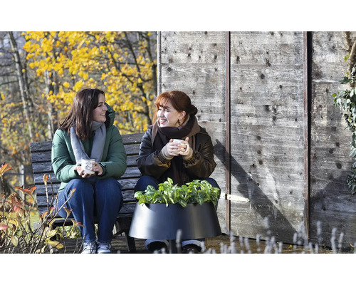 Zwei Frauen sitzen auf einer Holzbank im herbstlichen Garten neben einem schwarzen Pflanzgefäß mit grünen Pflanzen vor einer Holzwand.