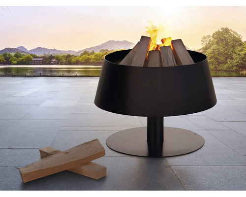 Moderne schwarze Feuerschale aus Metall mit brennendem Kaminholz auf einer Terrasse mit Steinplatten vor einem See und Bergen.