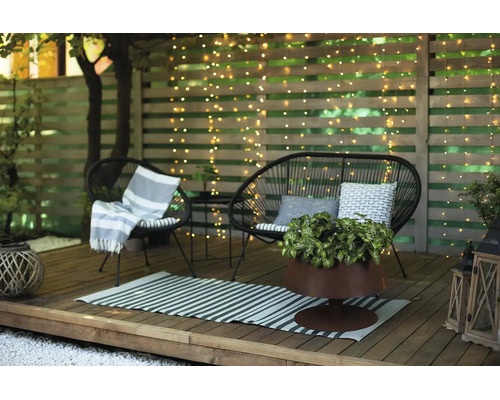Holzterrasse mit schwarzen Loungemöbeln, Lichterkette am Holzzaun, gestreiftem Teppich und Pflanzen.