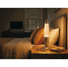 Lampe à lave dans une chambre sur une table de chevet en bois à côté d'un lit avec un éclairage chaleureux.