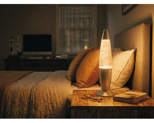 Lampe à lave dans une chambre sur une table de chevet en bois à côté d'un lit avec un éclairage chaleureux.