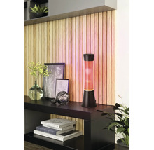 Lampe à lave moderne avec cire orange et socle noir sur un buffet devant un mur décoratif en tasseaux de bois.
