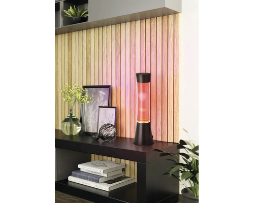 Lampe à lave moderne avec cire orange et socle noir sur un buffet devant un mur décoratif en tasseaux de bois.