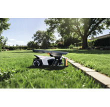 Robot tondeuse sous un abri sur une pelouse dans un jardin ensoleillé avec des arbres et une bordure en pierre.