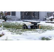 Robot tondeuse dans un garage sur une pelouse enneigée pendant une chute de neige devant une maison.