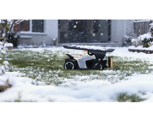 Robot tondeuse dans un garage sur une pelouse enneigée pendant une chute de neige devant une maison.