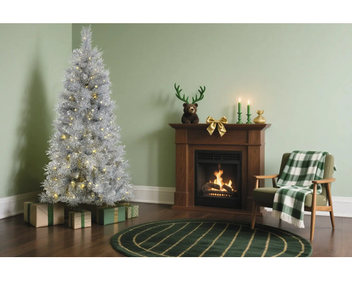 Silberner künstlicher Weihnachtsbaum mit Beleuchtung neben einem Kamin aus Holz, einem Sessel mit karierter Decke und grünem Teppich.