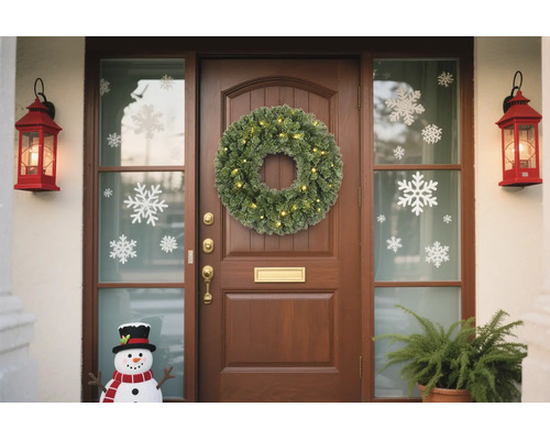 Hölzerne Haustür mit einem beleuchteten grünen Kranz, Schneeflocken-Fensterbildern, roten Laternen und einer Schneemannfigur.