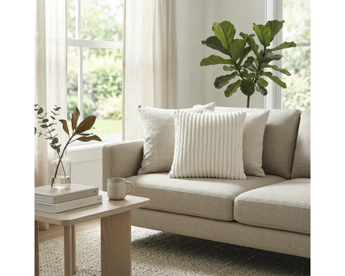 Helles Wohnzimmer mit beigem Stoffsofa, weissem Cord-Zierkissen und Holz-Beistelltisch. Grosse Zimmerpflanze und Fenster im Hintergrund.
