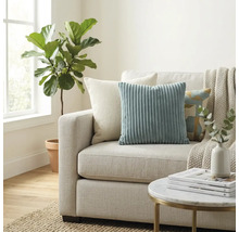 Helles Wohnzimmer mit Stoffsofa, Zierkissen aus Cord, einer Wohndecke, einem runden Beistelltisch und einer großen Zimmerpflanze neben dem Fenster.