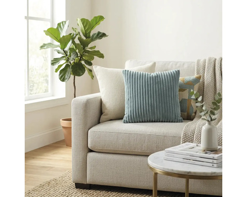 Helles Wohnzimmer mit Stoffsofa, Zierkissen aus Cord, einer Wohndecke, einem runden Beistelltisch und einer großen Zimmerpflanze neben dem Fenster.