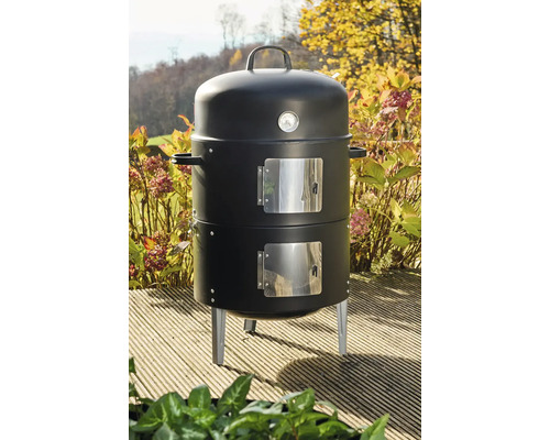 Schwarzer vertikaler Holzkohle-Smoker mit eingebautem Thermometer und zwei Türen auf einer hölzernen Terrasse im Garten.