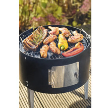 Runder schwarzer Holzkohlegrill mit gegrilltem Fleisch, Würstchen und Paprika auf einer Holzterrasse.