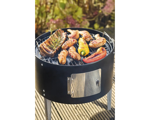 Runder schwarzer Holzkohlegrill mit gegrilltem Fleisch, Würstchen und Paprika auf einer Holzterrasse.