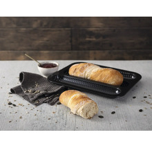 Schwarzes, gelochtes Baguette-Backblech aus Metall mit frisch gebackenem Brot in einer hellen, organisierten Küchenszene auf einem Holztisch.