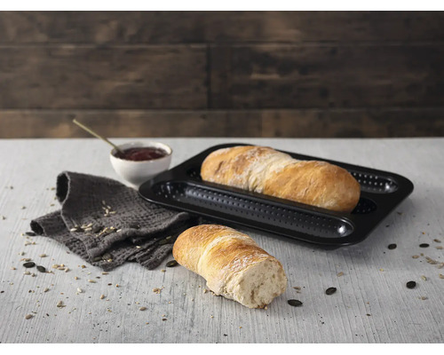 Schwarzes, gelochtes Baguette-Backblech aus Metall mit frisch gebackenem Brot in einer hellen, organisierten Küchenszene auf einem Holztisch.