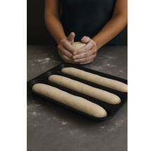 Schwarzes gelochtes Baguette-Backblech mit drei Teigrohlingen und eine Person, die im Hintergrund Teig formt.