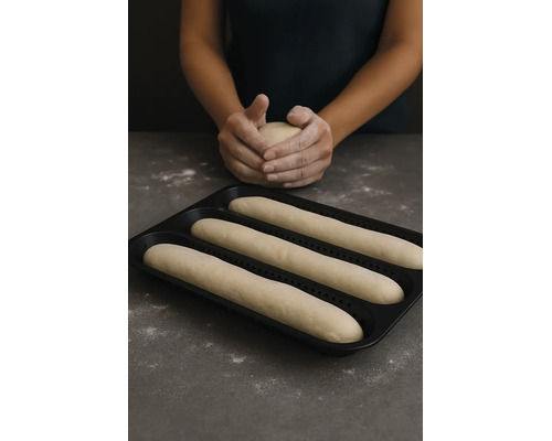Schwarzes gelochtes Baguette-Backblech mit drei Teigrohlingen und eine Person, die im Hintergrund Teig formt.