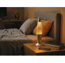 Lampe à lave à paillettes en métal sur une table de chevet en bois dans une chambre à coucher à côté d'un lit.