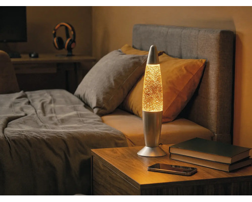 Lampe à lave à paillettes en métal sur une table de chevet en bois dans une chambre à coucher à côté d'un lit.