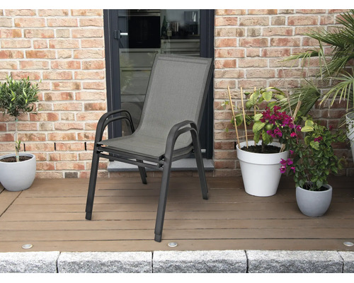 Stapelbarer Gartenstuhl mit Aluminiumgestell und Textilbespannung auf einer Terrasse mit Holzdeck und Pflanzen vor einer Ziegelwand.