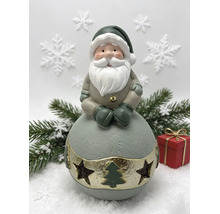 Figurine de Père Noël sur une boule avec découpes d'étoiles et de sapin, branches de sapin et cadeau en arrière-plan.