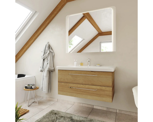 Salle de bains moderne avec meuble sous-vasque en chêne, lavabo blanc et miroir éclairé sous un toit en pente avec poutres en bois.