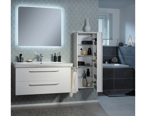 Salle de bains moderne avec meuble sous-lavabo blanc, miroir éclairé et armoire haute ouverte avec étagères en verre devant un mur tapissé.