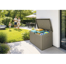 Coffre de jardin ouvert en plastique aspect bois sur terrasse, rempli de coussins et jouets, jardin ensoleillé avec une famille en arrière-plan.