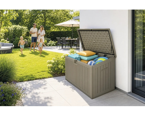 Coffre de jardin ouvert en plastique aspect bois sur terrasse, rempli de coussins et jouets, jardin ensoleillé avec une famille en arrière-plan.