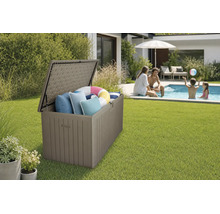 Coffre de rangement de jardin ouvert en plastique aspect bois sur une pelouse près d'une piscine, rempli de coussins et d'une bouée.
