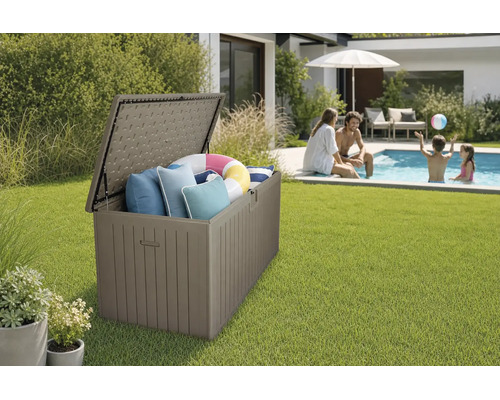 Coffre de rangement de jardin ouvert en plastique aspect bois sur une pelouse près d'une piscine, rempli de coussins et d'une bouée.