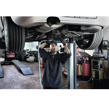 Mechaniker arbeitet in einer Werkstatt mit einem Druckluftwerkzeug am Unterboden eines Autos auf einer Hebebühne.