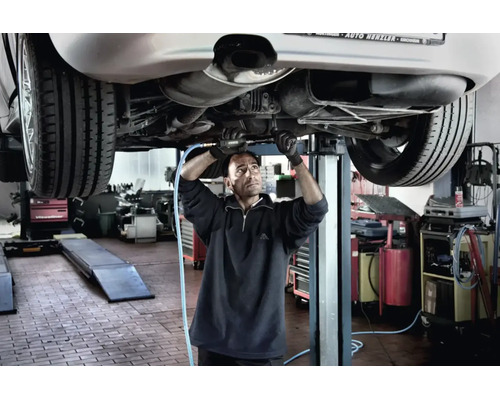 Mechaniker arbeitet in einer Werkstatt mit einem Druckluftwerkzeug am Unterboden eines Autos auf einer Hebebühne.